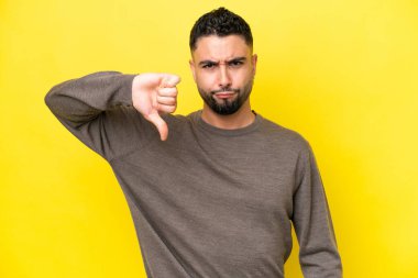 Sarı arka planda izole edilmiş genç Arap yakışıklı adam olumsuz ifadeyle baş parmağını aşağı gösteriyor.