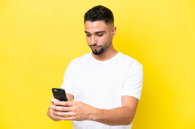 Genç Arap yakışıklı adam sarı arka planda izole edilmiş cep telefonuyla mesaj ya da e-posta gönderiyor.