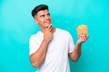 Young caucasian man catching french fries isolated on blue background looking up while smiling