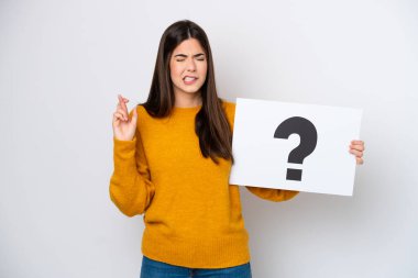 Young Brazilian woman isolated on white background holding a placard with question mark symbol with fingers crossing