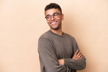 Young Brazilian man isolated on beige background with arms crossed and happy