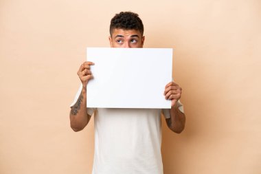 Genç Brezilyalı bir adam bej arka planda izole edilmiş boş bir levha tutuyor ve arkasında saklanıyor.