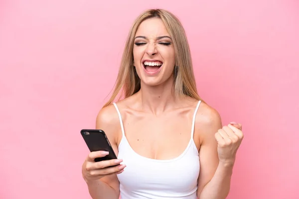 Pretty blonde woman isolated on pink background using mobile phone and doing victory gesture
