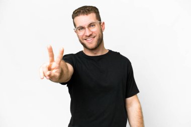 Genç yakışıklı sarışın adam izole edilmiş beyaz arka planda gülümsüyor ve zafer işareti gösteriyor.