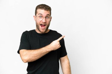 Yakışıklı sarışın, soyutlanmış beyaz bir arka plan. Şaşırmış ve işaret ediyor.