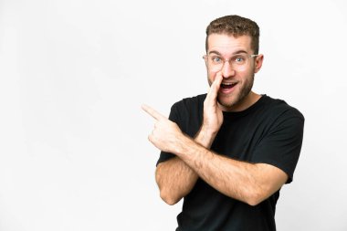 Young handsome blonde man over isolated white background pointing to the side to present a product and whispering something