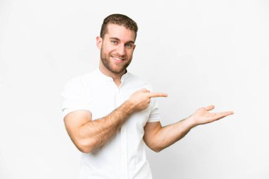 Young handsome blonde man over isolated white background holding copyspace imaginary on the palm to insert an ad