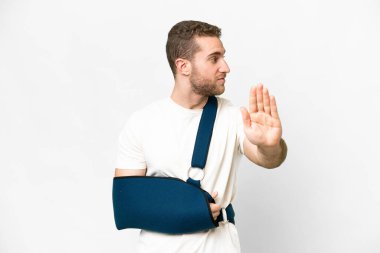 Young handsome blonde man with broken arm and wearing a sling over isolated white background making stop gesture and disappointed