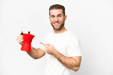Young handsome blonde man holding coffee pot over isolated white background and pointing it