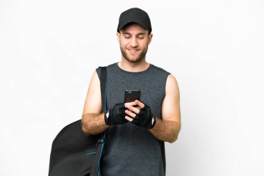 Young sport man with sport bag over isolated white background sending a message with the mobile