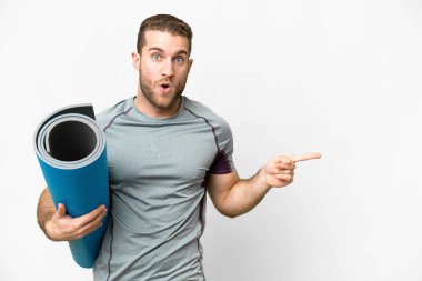 Young sport man going to yoga classes while holding a mat over isolated white background surprised and pointing side