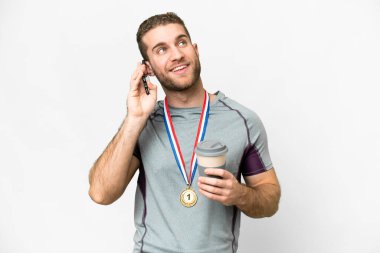 İzole edilmiş beyaz arka planda madalyaları olan yakışıklı sarışın genç bir adam. Elinde kahve ve cep telefonu var.