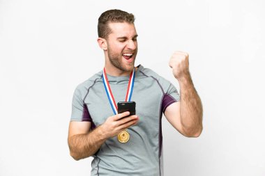 Young handsome blonde man with medals over isolated white background with phone in victory position