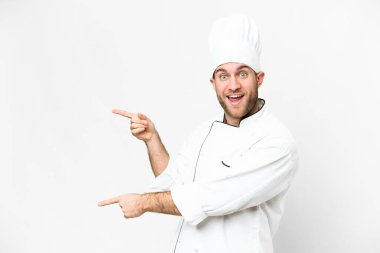 Young blonde man Chef over isolated white background surprised and pointing side