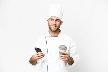 Young blonde man Chef over isolated white background holding coffee to take away and a mobile while thinking something