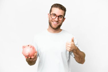 Young handsome blonde man holding a piggybank with thumbs up because something good has happened