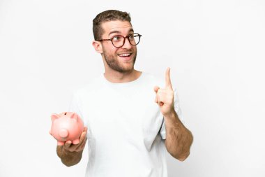 Young handsome blonde man holding a piggybank intending to realizes the solution while lifting a finger up