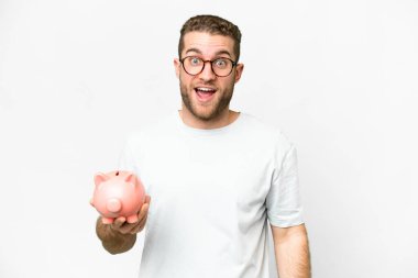 Young handsome blonde man holding a piggybank with surprise and shocked facial expression