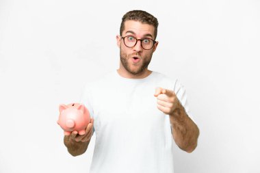 Young handsome blonde man holding a piggybank surprised and pointing front