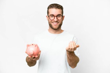 Young handsome blonde man holding a piggybank inviting to come with hand. Happy that you came