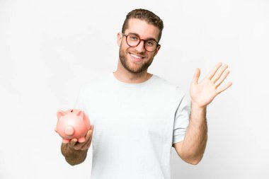 Young handsome blonde man holding a piggybank saluting with hand with happy expression