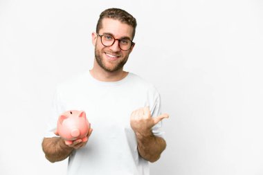 Young handsome blonde man holding a piggybank pointing to the side to present a product