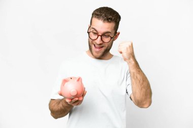 Young handsome blonde man holding a piggybank celebrating a victory