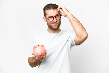 Young handsome blonde man holding a piggybank having doubts and with confuse face expression
