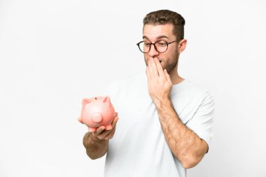 Young handsome blonde man holding a piggybank with surprise and shocked facial expression