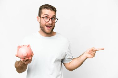 Young handsome blonde man holding a piggybank surprised and pointing finger to the side