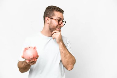 Young handsome blonde man holding a piggybank thinking an idea and looking side