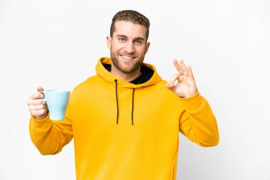 Young handsome blonde man holding cup of coffee over isolated background showing ok sign with fingers