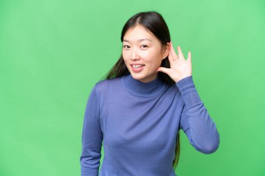 Young Asian woman over isolated chroma key background listening to something by putting hand on the ear