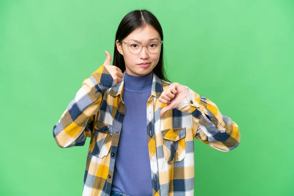 Young Asian woman over isolated chroma key background making good-bad sign. Undecided between yes or not