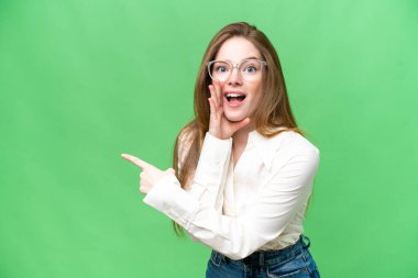 Young pretty woman over isolated chroma key background pointing to the side to present a product and whispering something