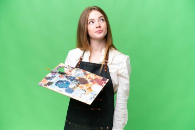 Young artist woman holding a palette over isolated chroma key background and looking up
