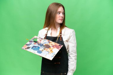 İzole edilmiş krom arka planda palet tutan genç sanatçı kadın yan tarafa bakıyor.