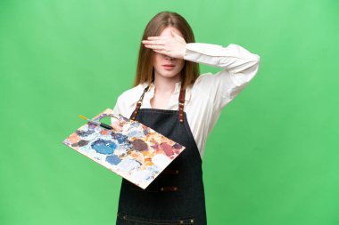 Young artist woman holding a palette over isolated chroma key background covering eyes by hands. Do not want to see something