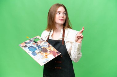 Young artist woman holding a palette over isolated chroma key background with fingers crossing and wishing the best