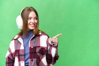 Young pretty woman wearing winter muffs over isolated chroma key background pointing finger to the side and presenting a product