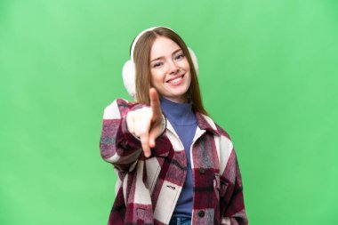 Young pretty woman wearing winter muffs over isolated chroma key background points finger at you with a confident expression