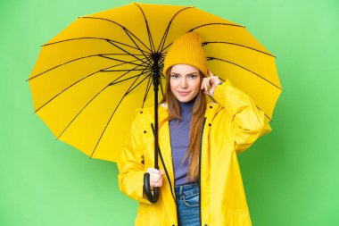 Young pretty woman with rainproof coat and umbrella over isolated chroma key background having doubts