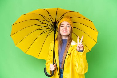 Young pretty woman with rainproof coat and umbrella over isolated chroma key background smiling and showing victory sign