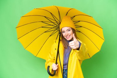 Young pretty woman with rainproof coat and umbrella over isolated chroma key background with thumbs up because something good has happened