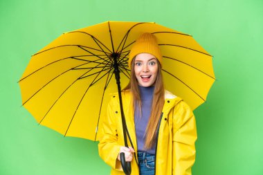 Young pretty woman with rainproof coat and umbrella over isolated chroma key background with surprise facial expression