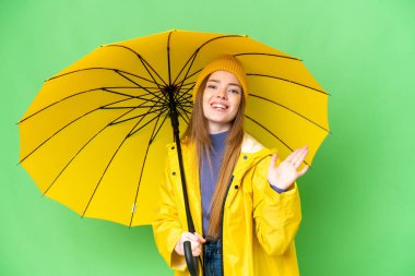 Young pretty woman with rainproof coat and umbrella over isolated chroma key background saluting with hand with happy expression
