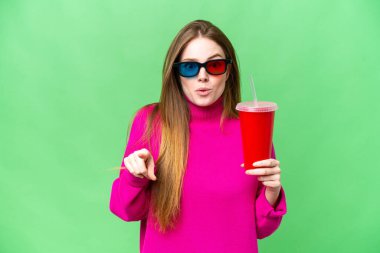 Young woman holding soda while watching a 3D movie surprised and pointing front