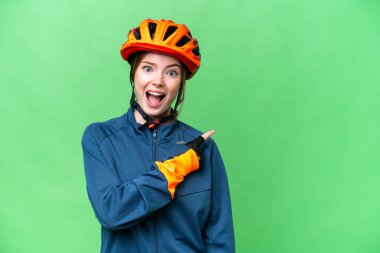 Young cyclist woman over isolated chroma key background surprised and pointing side