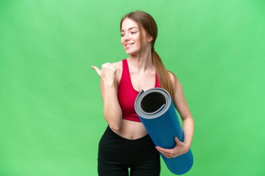 Genç sporcu kadın yoga derslerine giderken izole edilmiş krom anahtar arkaplanının üzerinde bir paspas tutuyor ve bir ürünü sunmak için yan tarafı işaret ediyor.