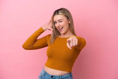 Pembe arka planda izole edilmiş genç beyaz kadın telefon hareketlerinde bulunuyor ve ön tarafı işaret ediyor.